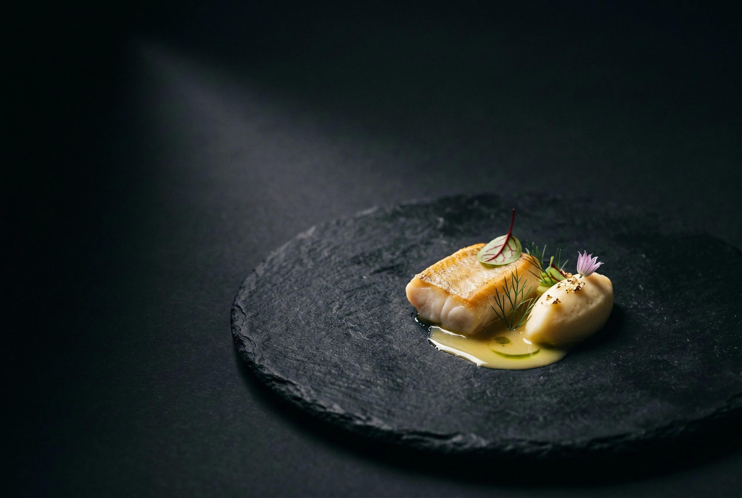 A composed tasting-menu course arranged on a dark stone plate, the dish occupying the lower-right third of the frame with expansive dark negative space above and to the left