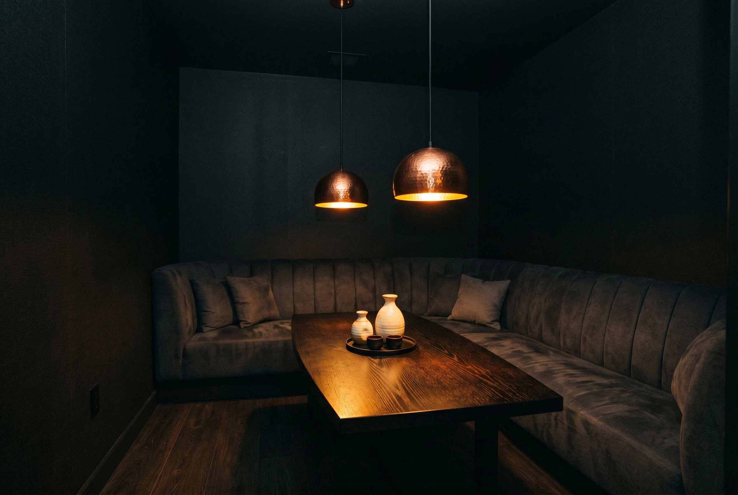 The private lounge interior at evening, a low banquette seat curving along one wall, sake vessels on the table surface, warm amber pendant lights casting pools of light over dark wood