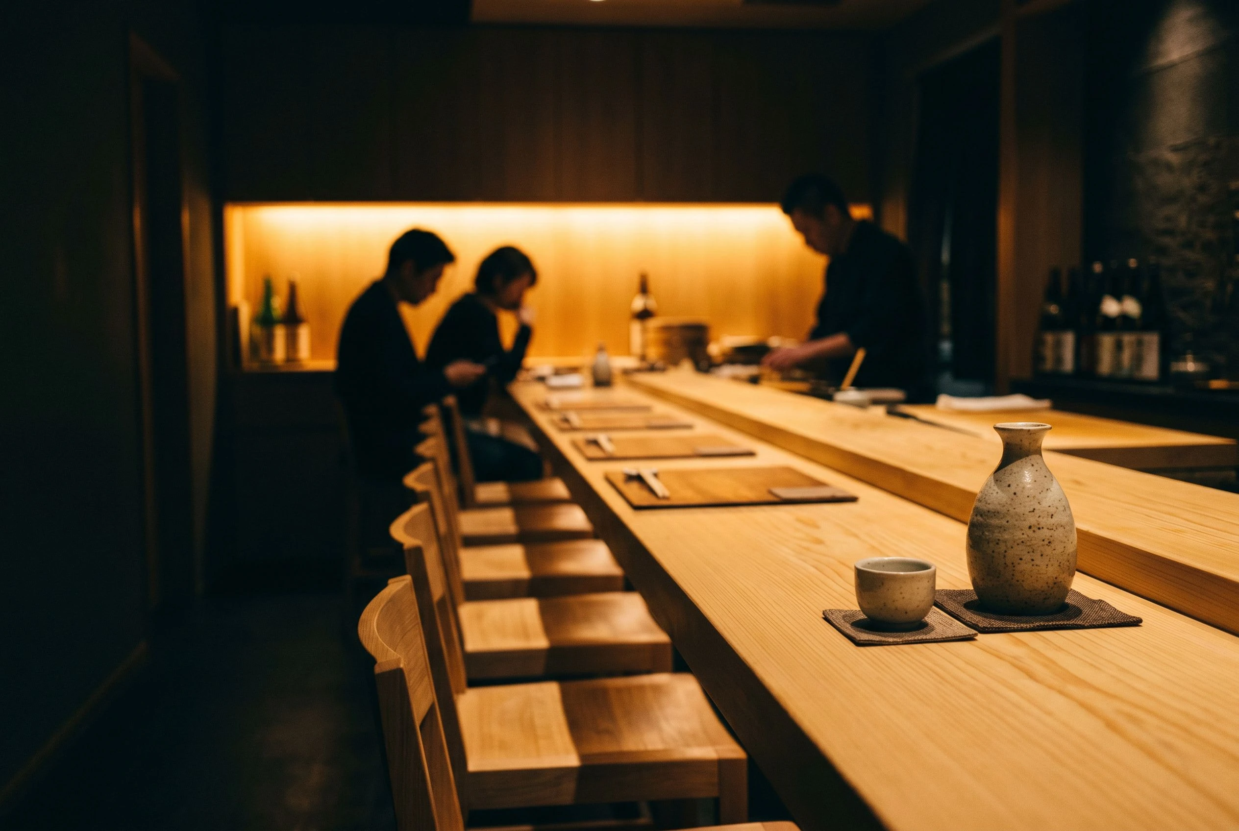 The omakase bar counter stretches across the frame with eight pale hinoki-wood seats, two guests seated mid-service in soft focus, warm amber backlight glowing behind the counter