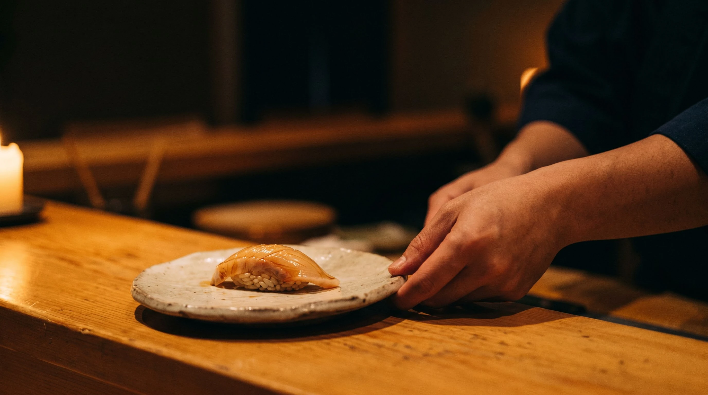 A single piece of nigiri rests on a pale ceramic dish at the omakase counter, illuminated by warm candlelight reflected in the lacquered wood surface, a chef's hands visible at the edge of the frame