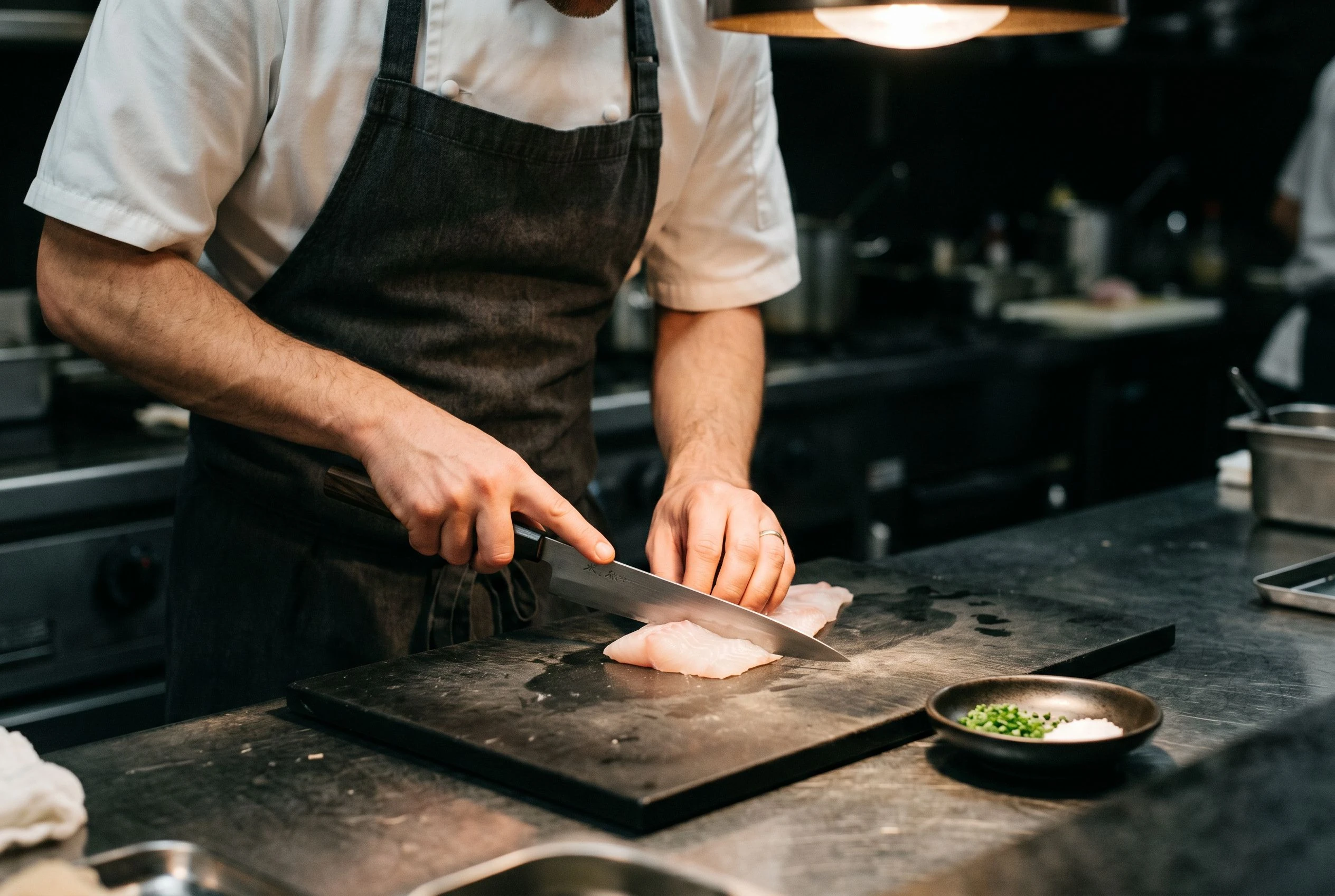 A chef's hands and forearms in sharp focus as they perform precise knife work on a fish portion at a preparation counter, the chef's face deliberately out of frame above the crop line