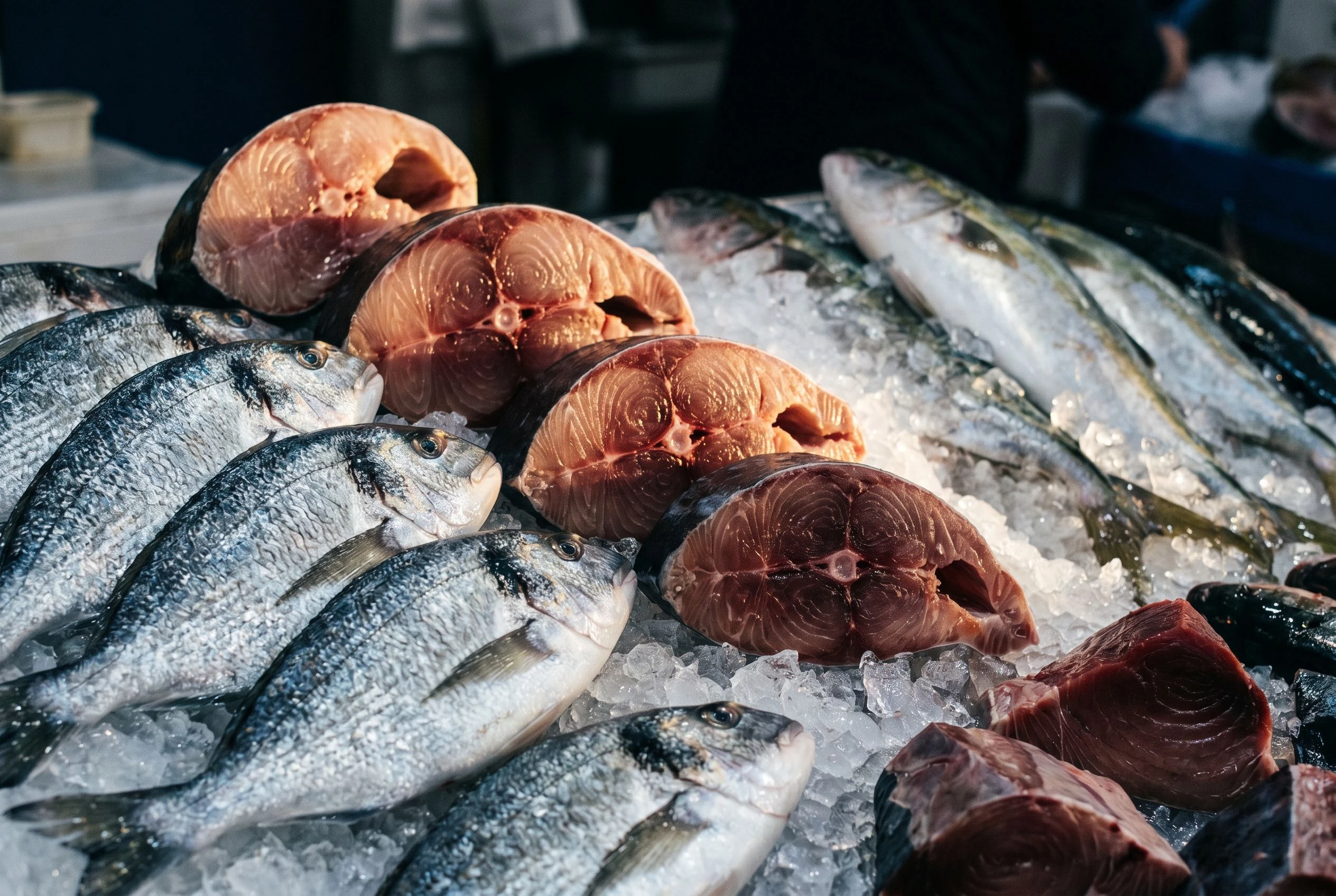 A close selection of whole fish and premium cuts gleaming with moisture at a fish market in early morning light, silver and gold tones dominant, ice crystals visible beneath