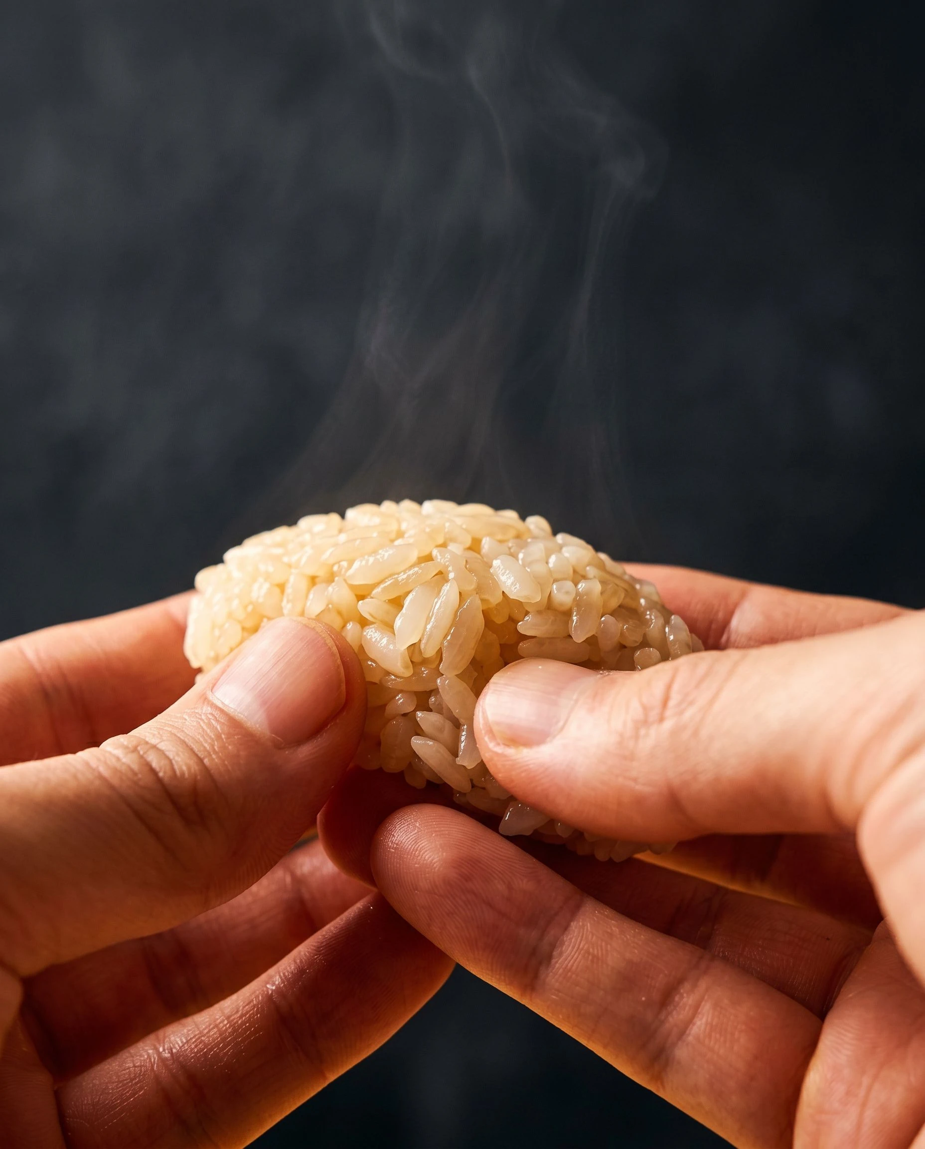 An extreme close-up of two hands shaping a portion of sushi rice, wisps of steam rising from the warm rice, the texture of each grain visible in warm amber light